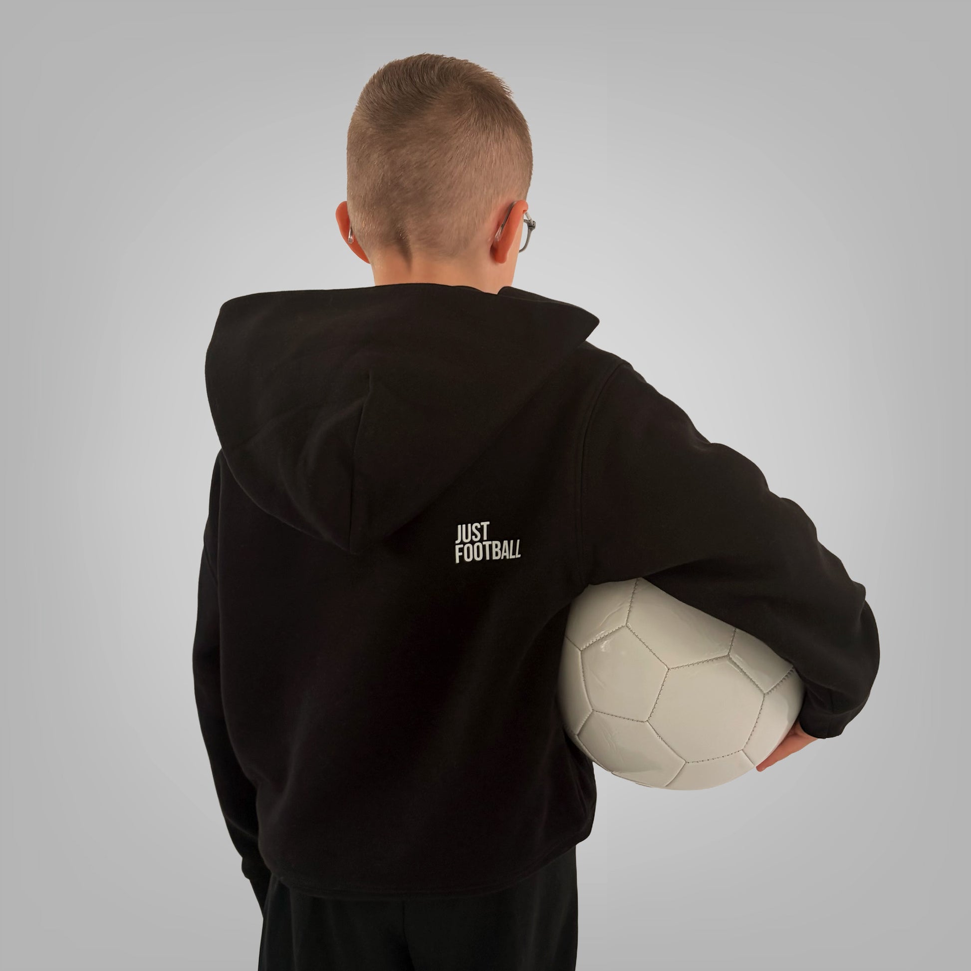 Back of black ONETWENTI Just Football hoodie worn by younger boy model with football