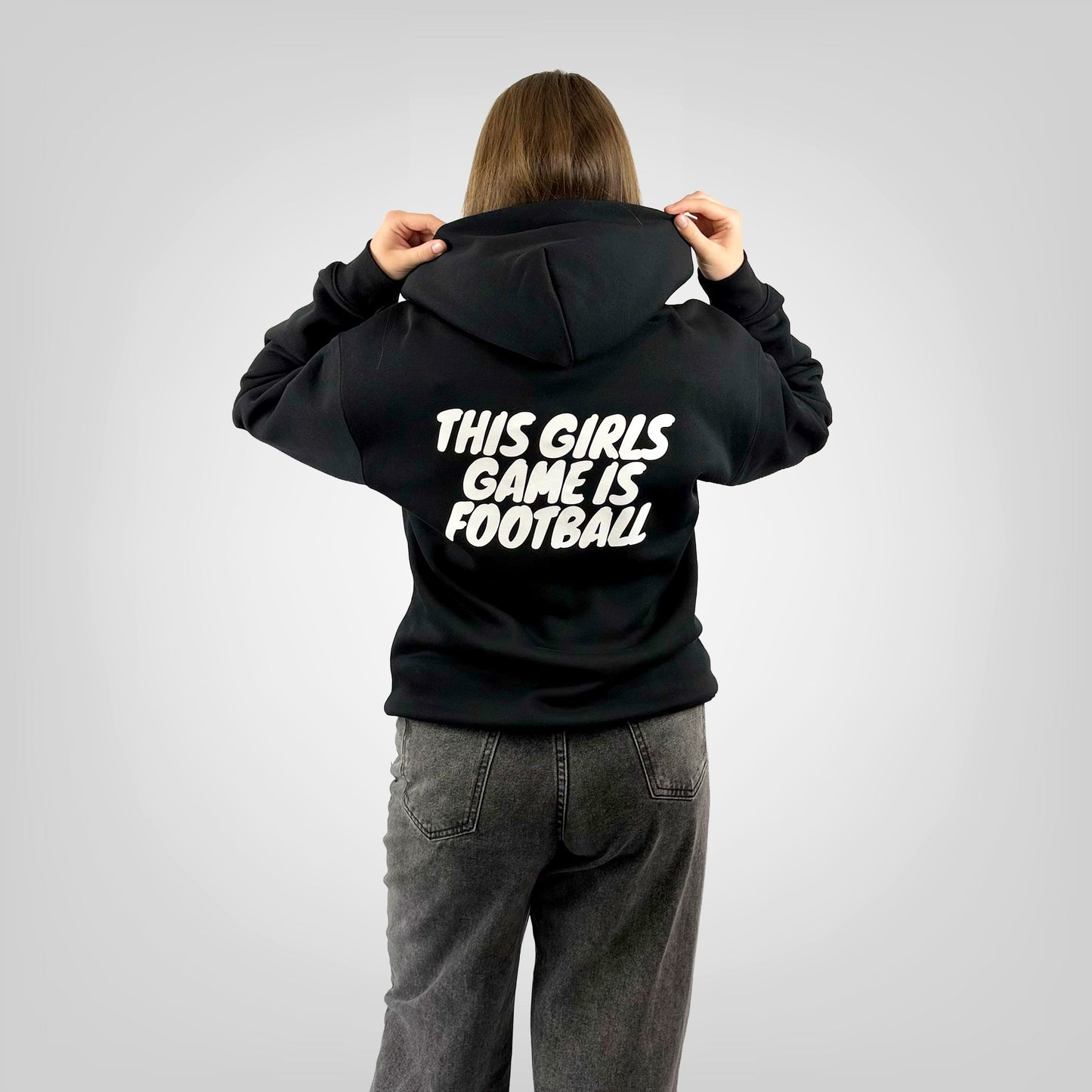 Back view of a female model wearing the ONETWENTI This Girls Game Is Football relaxed fit hoodie in black.